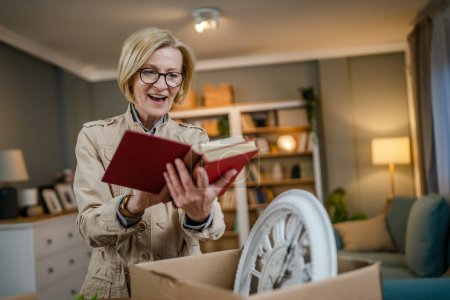 Photo for One senior woman pensioner grandmother moving in new apartment taking her stuff belongings and plants in or out of boxes packing or unpacking real person hold books copy space - Royalty Free Image