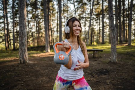 Foto de One woman adult caucasian female athlete using kettlebell girya weight during training in the forest park woods in summer day with headphones happy smile brunette health and fitness concept copy space - Imagen libre de derechos