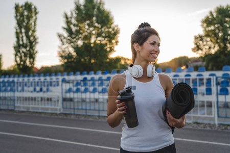 Téléchargez les photos : One woman adult caucasian female athlete standing on stadium on running track with supplement shaker and yoga mat with headphones ready for training outdoor in summer evening real people copy space - en image libre de droit