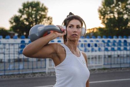 Téléchargez les photos : One woman adult caucasian female athlete training with kettlebell girya weight in stadium on track in summer evening or morning with headphones happy brunette health and fitness concept copy space - en image libre de droit