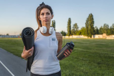 Téléchargez les photos : One woman adult caucasian female athlete standing on stadium on running track with supplement shaker and yoga mat with headphones ready for training outdoor in summer evening real people copy space - en image libre de droit