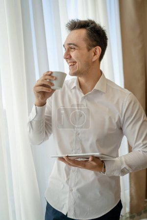 Foto de One man adult caucasian male standing by the window at home office or hotel room with cup of coffee and digital tablet taking a brake from work and preparing real people copy space wear white shirt - Imagen libre de derechos