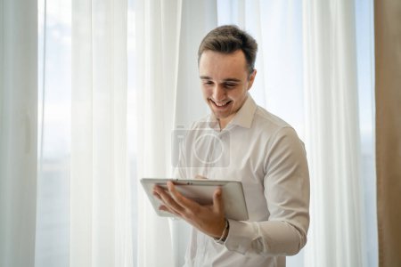 Téléchargez les photos : One man adult caucasian male standing by the window at home office or hotel room with cup of coffee and digital tablet taking a brake from work and preparing real people copy space wear white shirt - en image libre de droit