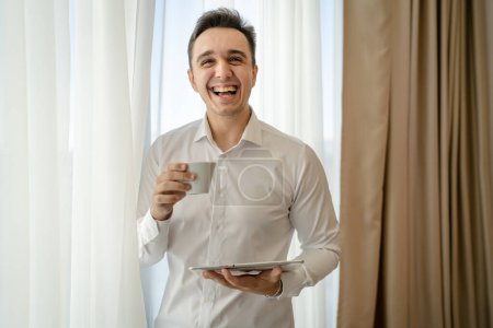 Foto de One man adult caucasian male standing by the window at home office or hotel room with cup of coffee and digital tablet taking a brake from work and preparing real people copy space wear white shirt - Imagen libre de derechos