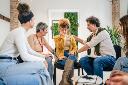 Photo for A group of people sitting in a circle with two therapists and a woman in the center discussing and providing emotional support for her The focus is on the woman but everyone is engaged in conversation - Royalty Free Image