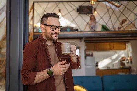 Photo for Man caucasian 40 years old male wear a shirt stands on a balcony holding a cup of tea or coffee smiling happy He is enjoying a peaceful moment in his daily morning routine or on a vacation copy space - Royalty Free Image