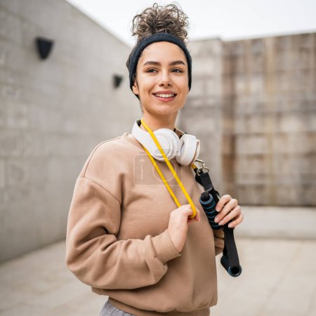 Téléchargez les photos : Portrait d'une jeune femme caucasienne adulte debout en plein air devant un mur en béton moderne avec écouteurs confiants avec bande de résistance en caoutchouc tubes espace de copie - en image libre de droit