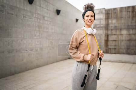 Photo for Portrait of one young adult caucasian woman stand outdoor in front of modern concrete wall with headphones confident with rubber resistance band tubes copy space - Royalty Free Image