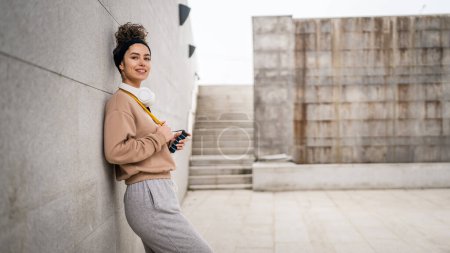 Téléchargez les photos : Portrait d'une jeune femme caucasienne adulte debout en plein air devant un mur en béton moderne avec écouteurs confiants avec bande de résistance en caoutchouc tubes espace de copie - en image libre de droit