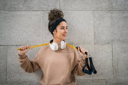 Photo for Portrait of one young adult caucasian woman stand outdoor in front of modern concrete wall with headphones confident with rubber resistance band tubes copy space - Royalty Free Image