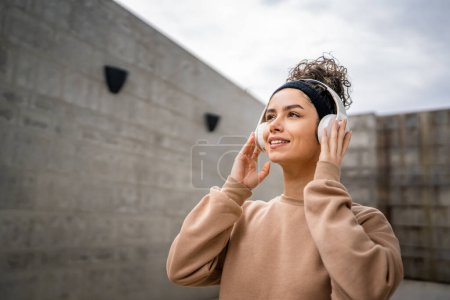 Foto de Retrato de una joven mujer caucásica adulta parada al aire libre frente a la pared de hormigón moderno con auriculares en la cabeza escuchar música confiada - Imagen libre de derechos
