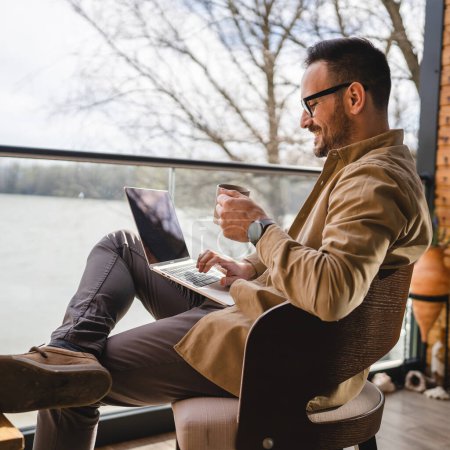 Photo for One man adult male 40 years old sit on a terrace balcony with a cup of coffee use laptop computer entrepreneur or freelancer on vacation morning routine real person copy space - Royalty Free Image