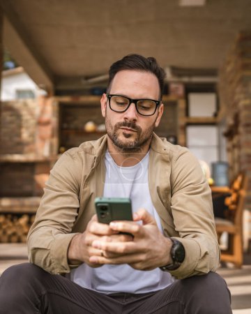 Téléchargez les photos : Un homme adulte 40 ans homme caucasien est assis devant sa maison utiliser smartphone pour lire des messages ou des nouvelles en profondeur dans la pensée inquiet ou préoccupé penser espace de copie personne réelle - en image libre de droit