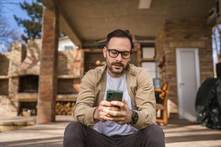 Téléchargez les photos : Un homme adulte 40 ans homme caucasien est assis devant sa maison utiliser smartphone pour lire des messages ou des nouvelles en profondeur dans la pensée inquiet ou préoccupé penser espace de copie personne réelle - en image libre de droit
