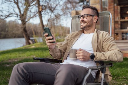 Foto de Un hombre adulto de 40 años caucásico macho sentarse frente a su casa en vacaciones hablar en su teléfono inteligente llamada de teléfono móvil sonrisa feliz durante la conversación persona real copiar espacio - Imagen libre de derechos