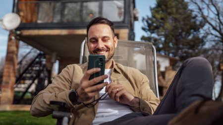 Photo for One adult man 40 years old caucasian male sit in front of his house on vacation talk on his smartphone mobile phone call happy smile during the conversation real person copy space - Royalty Free Image
