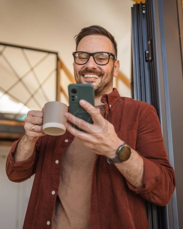Téléchargez les photos : Un homme adulte caucasien mâle debout sur le balcon utiliser son smartphone sourire heureux tenant smartphone dans une main et debout près de la messagerie de porte ou en utilisant un réseau social en ligne sur Internet - en image libre de droit