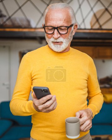Foto de Un hombre mayor con un soporte de barba en el balcón sostiene un teléfono móvil y una taza de café rutina diaria de la mañana espacio de copia en persona real leer o usar el teléfono inteligente Internet navegar por la red social - Imagen libre de derechos