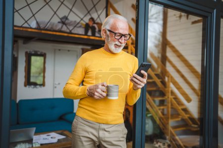 Photo for One senior man with a beard stand on the balcony hold a mobile phone and cup of coffee happy smile daily morning routine real person copy space read or use smartphone internet browse social network - Royalty Free Image