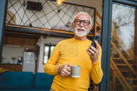 Téléchargez les photos : Un homme âgé avec un stand de barbe sur le balcon tenir un téléphone mobile et une tasse de café sourire heureux tous les jours matin routine personne réelle copier l'espace lu ou utiliser smartphone Internet naviguer sur le réseau social - en image libre de droit