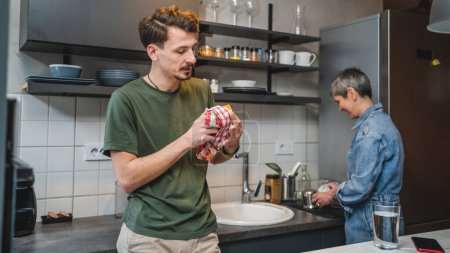 Photo for Adult Caucasian man is standing in the kitchen hold a cup and cloth while his mother take care of the dishes engaged in a conversation creating a special moment of connection and bond work together - Royalty Free Image