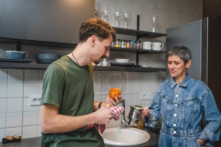 adult Caucasian man is standing in the kitchen hold a cup and cloth while his mother take care of the dishes engaged in a conversation creating a special moment of connection and bond work together