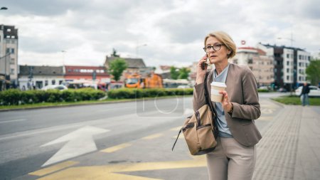 Photo for One woman caucasian mature blonde female with eyeglasses stand at bus city stop in day use smartphone mobile phone making a call talk while wait for the drive real person copy space late for work - Royalty Free Image