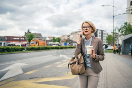 Téléchargez les photos : Une femme caucasienne mature blonde femme avec des lunettes se tenir à l'arrêt de bus ville dans la journée utiliser smartphone téléphone mobile faire un appel parler en attendant recevoir de mauvaises nouvelles - en image libre de droit