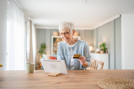Téléchargez les photos : Une femme âgée mature grand-mère s'asseoir à la maison utiliser carte de crédit ou de débit pour les achats en ligne parcourir les magasins en ligne acheter des choses vraies personnes copier l'espace - en image libre de droit