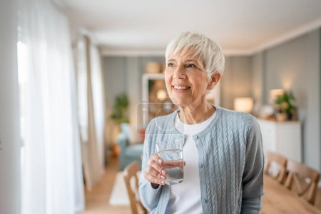 Téléchargez les photos : Gros plan portrait d'une femme âgée aux cheveux courts heureux sourire positif émotion copie espace debout à la maison intérieure grise cheveux blancs tenir verre d'eau - en image libre de droit