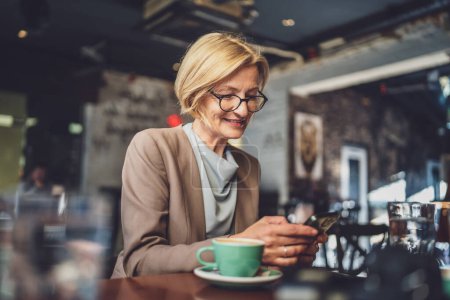 Photo for One stylish mature blonde Caucasian woman wearing eyeglasses sit comfortably in a cafe texting and browsing the internet on her smartphone modern lifestyle digital interactions of mature individuals - Royalty Free Image
