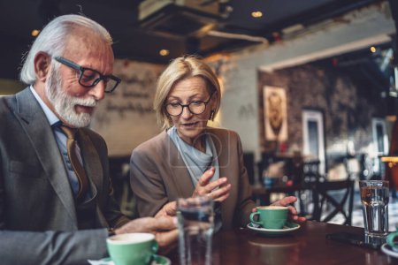 Photo for Senior man with a distinguished beard and a mature blonde woman whether as husband and wife business partners or friends share a joyful moment at a cafe look at photos or connect with others - Royalty Free Image