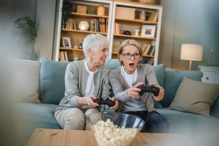 Photo for Two senior women caucasian friends or sisters happy old siblings pensioner playing video game console using joystick or controllers while sitting at home real people family leisure concept copy space - Royalty Free Image