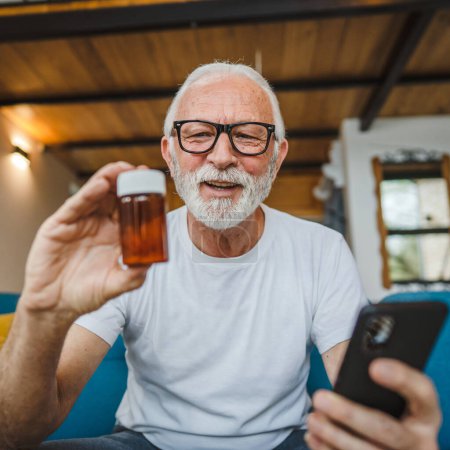 Photo for One senior man sitting at home on sofa bed holding bottle of medicine pills medicament or supplement and smartphone read label instructions of drug tablet painkillers or vitamin ready to take medicine - Royalty Free Image