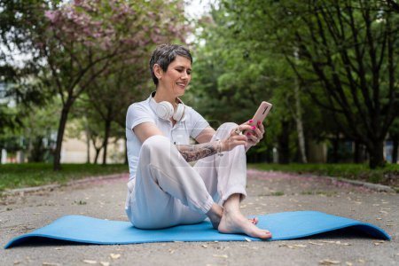 Téléchargez les photos : Une femme caucasienne mature âgée assise en plein air sur un tapis de yoga utilise un téléphone mobile pour une formation en ligne ou une pratique de méditation guidée. - en image libre de droit