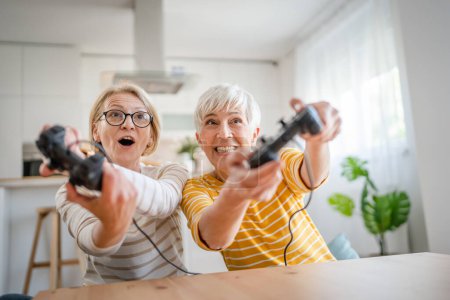 Photo for Two senior women caucasian friends or sisters happy old siblings pensioner playing video game console using joystick or controllers while sitting at home real people family leisure concept copy space - Royalty Free Image