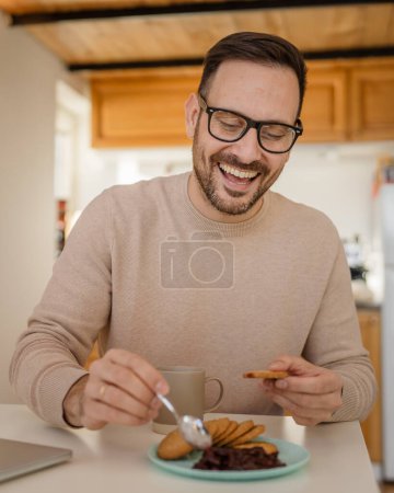 Foto de Un hombre adulto caucásico viejo macho sentarse en una mesa en casa poniendo mermelada en una galleta preparándose para desayunar disfrutar de aperitivo relajado en su hogar ambiente persona real copiar espacio rutina diaria - Imagen libre de derechos