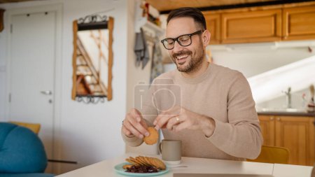 Téléchargez les photos : Un homme adulte caucasien vieux mâle assis à une table à la maison mettre de la confiture sur un biscuit se préparant à manger le petit déjeuner profiter d'une collation détendue dans son environnement à la maison personne réelle copier l'espace routine quotidienne - en image libre de droit
