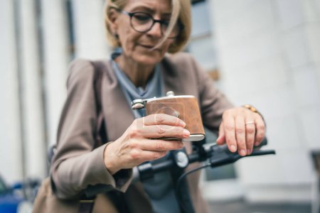 Photo for One woman serious mature caucasian female businesswoman stand outdoor with electric kick scooter hold flask drinking desperate depressed hard life concept alcohol abuse alcoholism selective focus - Royalty Free Image