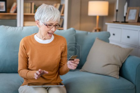 Téléchargez les photos : Une femme âgée mature grand-mère s'asseoir à la maison utiliser carte de crédit ou de débit pour les achats en ligne parcourir les magasins en ligne acheter des choses sur la vente à prix réduit personnes réelles copier l'espace utiliser une tablette numérique - en image libre de droit