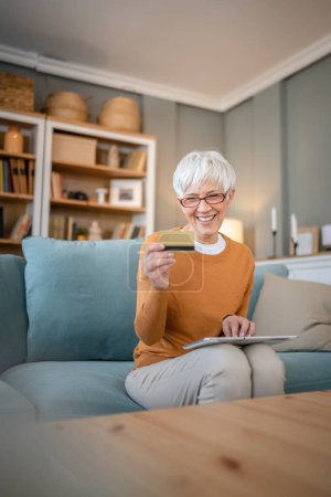Téléchargez les photos : Une femme âgée mature grand-mère s'asseoir à la maison utiliser carte de crédit ou de débit pour les achats en ligne parcourir les magasins en ligne acheter des choses sur la vente à prix réduit personnes réelles copier l'espace utiliser une tablette numérique - en image libre de droit