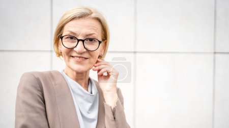 Photo for Portrait of one caucasian mature woman female businesswoman blonde hair and eyeglasses stand confident outdoor in front of white wall of modern building in day copy space - Royalty Free Image
