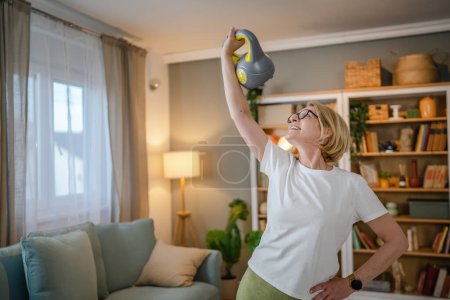 Foto de Una mujer rubia caucásica madura entrenando con kettlebell girya campana rusa en casa ejercicio femenino en su apartamento concepto de estilo de vida saludable - Imagen libre de derechos