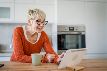 Photo for One woman mature blonde caucasian senior female at home use digital tablet for video call online internet chat talk on app real person copy space - Royalty Free Image