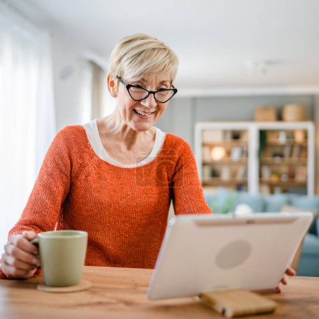 Photo for One woman mature blonde caucasian senior female at home use digital tablet for video call online internet chat talk on app real person copy space hold cup of coffee - Royalty Free Image