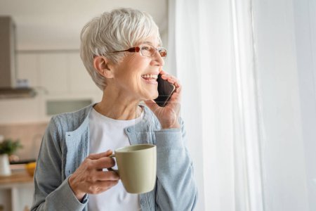 Eine ältere Seniorin steht zu Hause am Fenster und telefoniert mit dem Smartphone, echte Menschen stehen im hellen Raum, kurze graue Haare tragen Pullover und Brille.