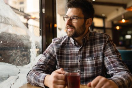 Foto de Adult caucasian man sitting alone at cafe front view of one male with beard and eyeglasses holding a cup of tea real people copy space - Imagen libre de derechos