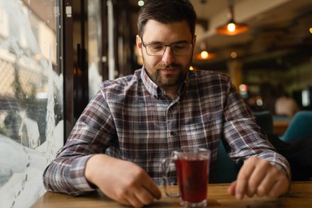 Téléchargez les photos : Adult caucasian man sitting alone at cafe front view of one male with beard and eyeglasses holding a cup of tea real people copy space - en image libre de droit