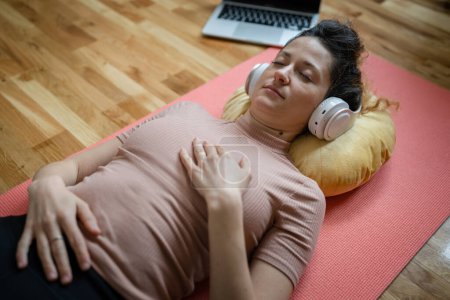 Téléchargez les photos : Une femme adulte caucasienne millénium en utilisant des écouteurs pour la méditation guidée en ligne pratiquant le yoga pleine conscience avec les yeux fermés sur le sol à la maison personnes réelles espace de copie concept d'auto-soins - en image libre de droit
