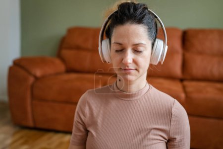 Téléchargez les photos : Une femme adulte caucasienne millénium en utilisant des écouteurs pour la méditation guidée en ligne pratiquant le yoga pleine conscience avec les yeux fermés sur le sol à la maison personnes réelles espace de copie concept d'auto-soins - en image libre de droit